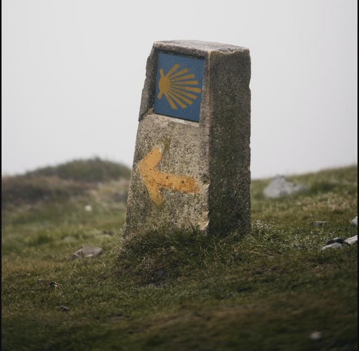 A stone trail marker with a blue and yellow Camino de Santiago shell and arrow painted on it
